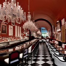 Load image into Gallery viewer, Elegant bar red walls vaulted ceiling grand crystal chandeliers black and white checkered flooring. white bar stools and chairs with black trim