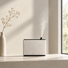 Load image into Gallery viewer, SMI_100- small space motion sensor scent diffuser in white with scent coming from the top, in a minimalist modern setting with a vase and flower on the left and window on the right