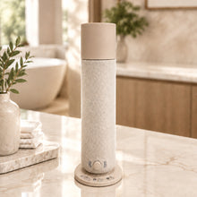 Load image into Gallery viewer, SMI 800 Scent diffuser tower on tan marble counter in modern upscale bathroom. vase on left, bath in background, vase on counter with green leaves in background
